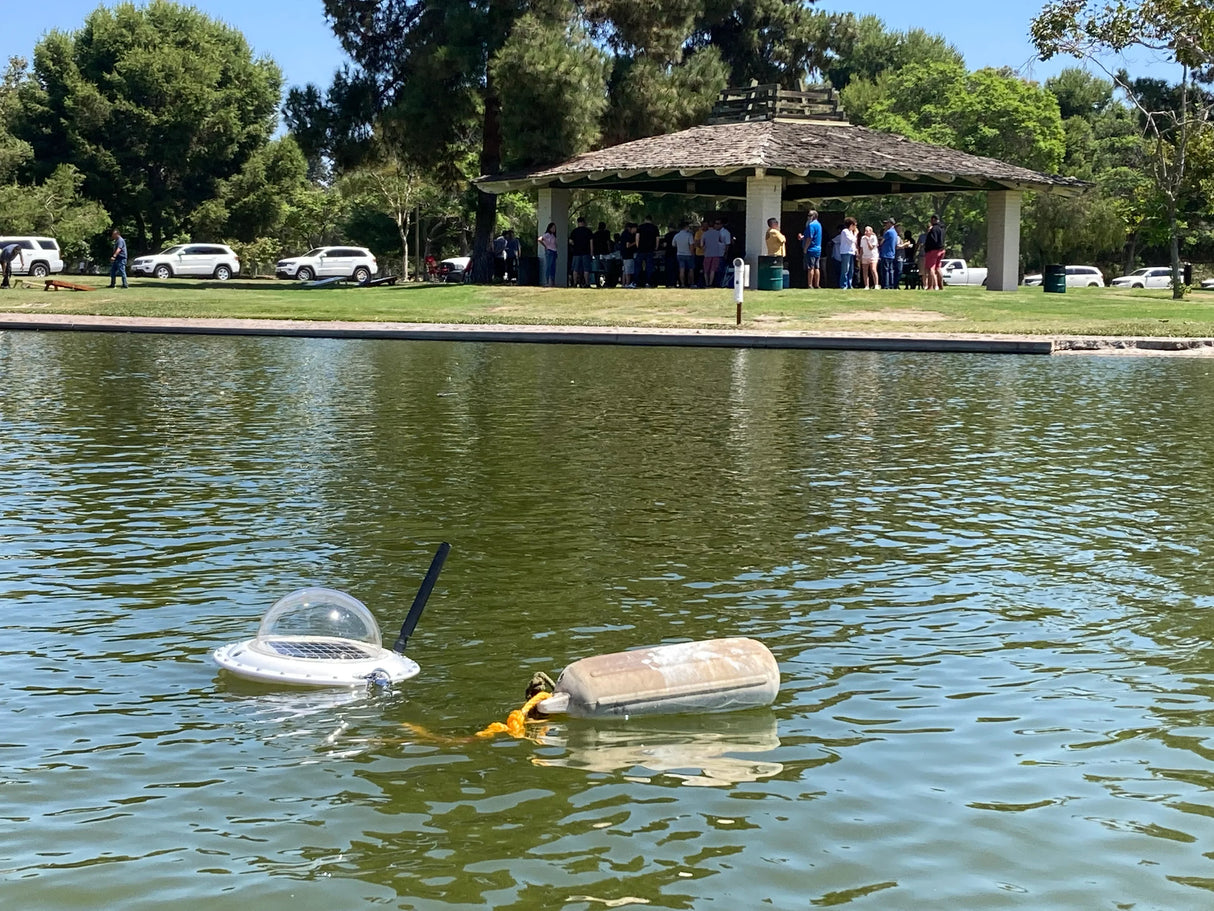 Lake Pulse water testing device floating on a lake near a park pavilion with people gathered.