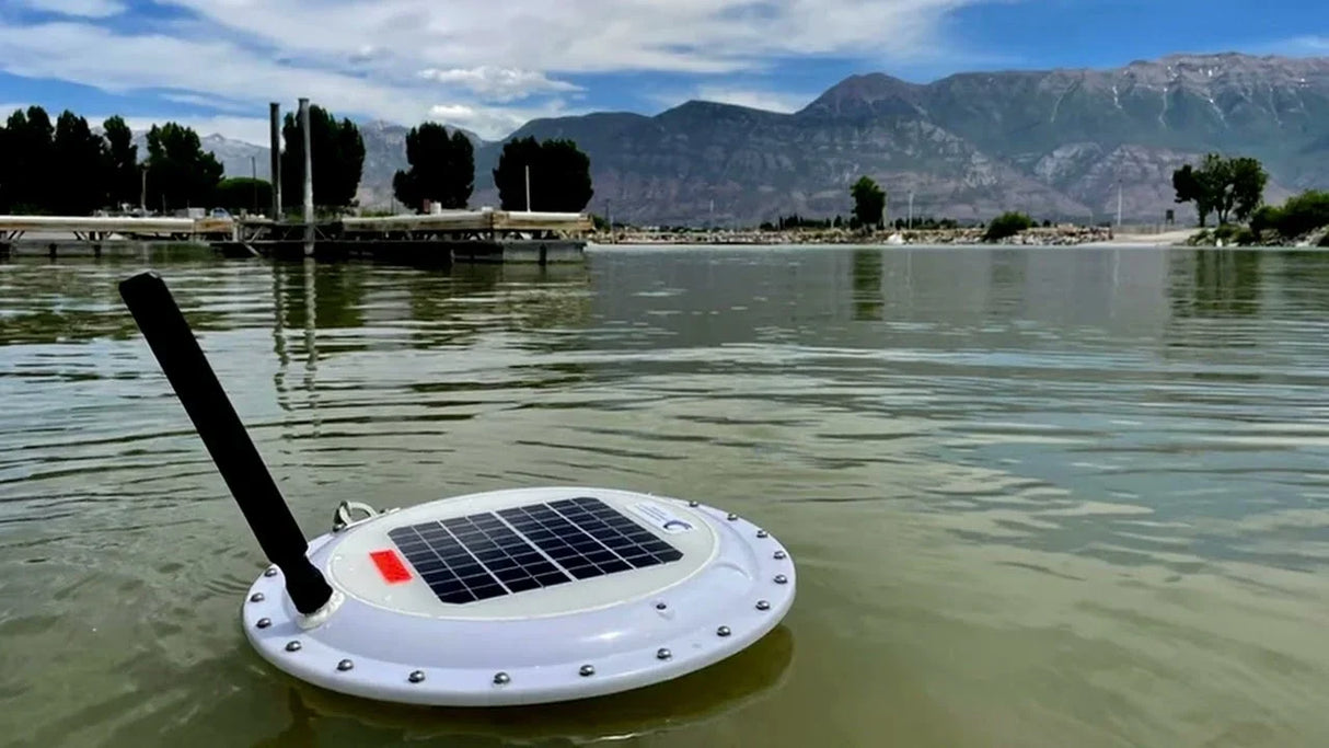 Solar-powered lake water monitoring device floating near shore, mountains in background
