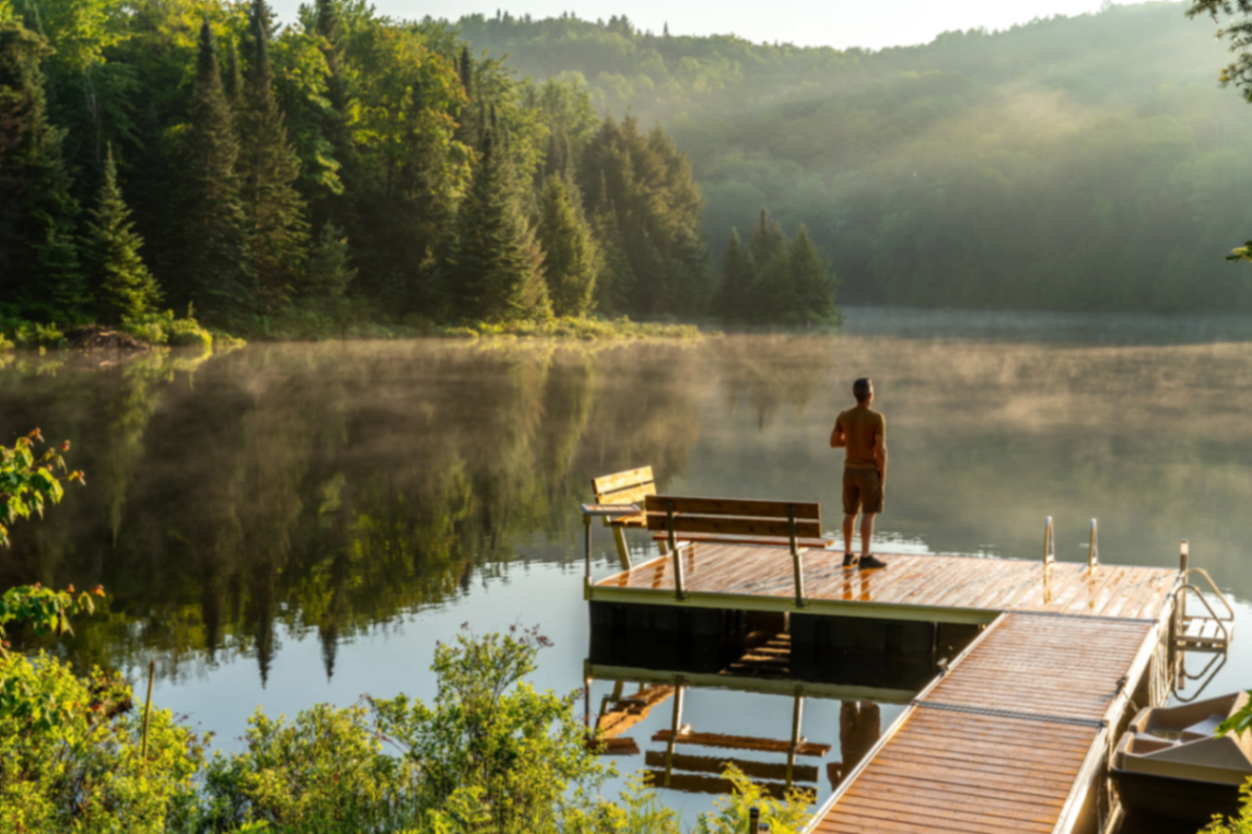A person standing on a wooden dock overlooking a calm forested lake in morning light